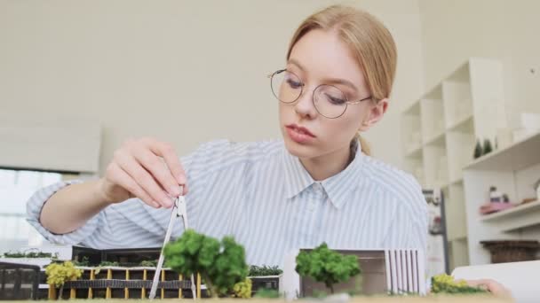 Улыбающаяся Молодая Блондинка Архитектор Очках Обозначает Модель Дома Сидя Современном — стоковое видео
