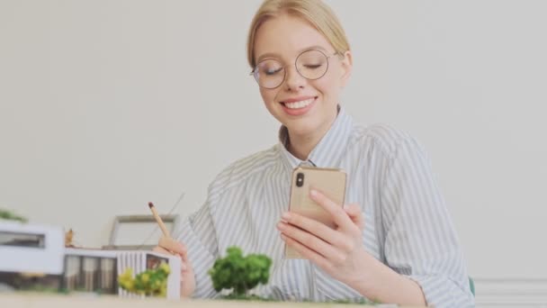 現代的なオフィスに座っている間 積極的な若いブロンドの女性建築家が眼鏡をかけて彼女のスマートフォンを使って何かを書いています — ストック動画