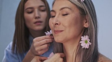 Genç bir makyöz, yaşlı ve mutlu bir kadın için güzellik salonundaki fotoğraf çekimi sırasında pembe çiçekleri saç stiline uyguluyor.