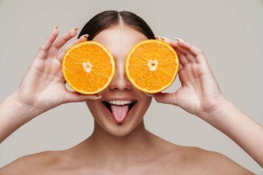 Gri arka planda portakalla eğlenirken gülümseyen, güzel, üstsüz bir kadın görüntüsü.