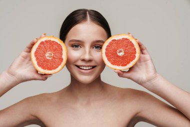 Gri arka planda izole edilmiş greyfurtla poz verirken gülümseyen neşeli, üstsüz kadın görüntüsü