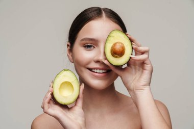 Gri arka planda izole edilmiş avokadoyla poz verirken gülümseyen neşeli, üstsüz kadın görüntüsü