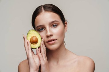 Gri arkaplanda, kamerada avokadoyla poz veren güzel üstsüz bir kadın resmi.