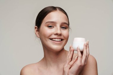 Yüzüne krem süren ve gri arka planda izole bir şekilde gülümseyen mutlu genç kadın görüntüsü