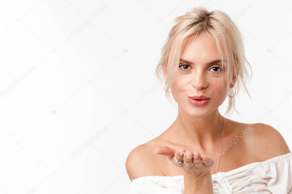 Image of joyful blonde woman in earrings blowing air kiss at camera isolated over white background