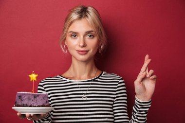 İyi şans dilemek için pasta tutan ve parmaklarını çapraz tutan genç bir kadın resmi kırmızı arka planda izole edilmiş.