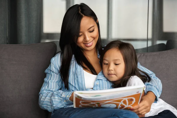 照片中 快乐的亚洲孕妇和她的小女儿坐在沙发上 抱着看书 — 图库照片