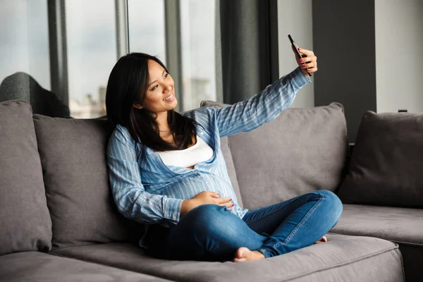 亚洲孕妇坐在沙发上 在手机上自拍的照片 — 图库照片