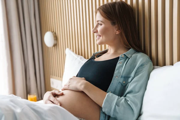 幸せな妊娠中の女性の写真付き赤い髪笑顔と彼女の腹をペットしながら 日当たりの良い部屋のベッドに横たわって — ストック写真