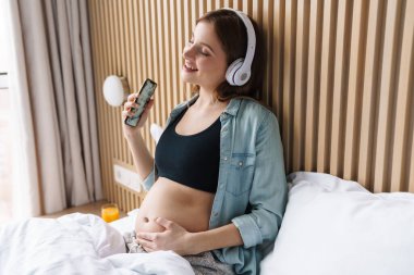 Güneşli odada yatakta şarkı söylerken cep telefonu ve kablosuz kulaklık kullanan mutlu hamile kadının fotoğrafı.