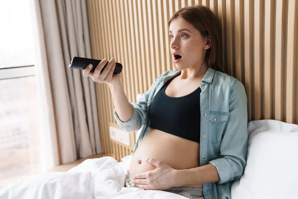 Foto Mulher Grávida Chocada Usando Controle Remoto Enquanto Assiste Cama — Fotografia de Stock