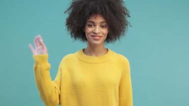 Kıvırcık saçlı mutlu Afro-Amerikan bir kadın elini sallayarak stüdyodaki mavi duvarın arkasında tek başına duran kameraya selam veriyor.