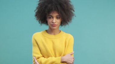 Kıvırcık saçlı sakin bir Afro-Amerikalı kadın kameraya bakıyor ve elleri çaprazlanmış bir şekilde stüdyoda mavi duvarın arkasında duruyor.