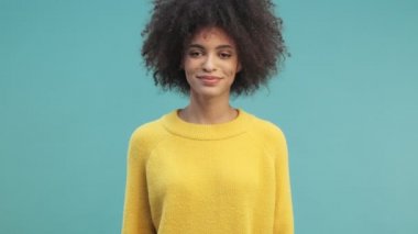 Kıvırcık saçlı, güzel, Afro-Amerikan bir kadın gülüyor. Stüdyoda mavi duvarın arkasında tek başına duruyor.