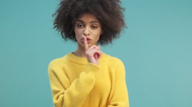 Kıvırcık saçlı hoş, Afro-Amerikan bir kadın stüdyoda, mavi duvarın ardından izole edilmiş bir sessizlik jesti yapıyor.