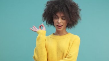 Kıvırcık saçlı başarılı bir Afro-Amerikan kadını stüdyoda mavi duvar arkasında izole edilmiş bir jest yapıyor.