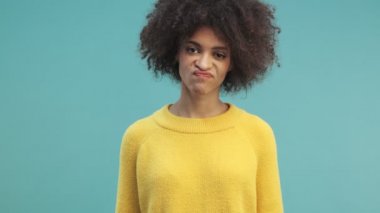 Kıvırcık saçlı, hoşnutsuz bir Afro-Amerikan kadın stüdyoda mavi duvarın arkasında izole bir jest yapıyor.
