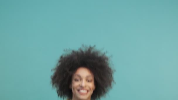 一位有着卷发的快乐的年轻的非洲裔美国女人在工作室里跳着 高兴地从蓝色的墙壁背景中跳了出来 — 图库视频影像