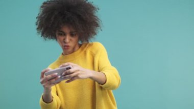 Kıvırcık saçlı mutlu bir Afrikalı Amerikalı kadın akıllı telefonuyla oyun oynuyor. Stüdyoda, mavi duvar arkasında onu yatay olarak izole ediyor.