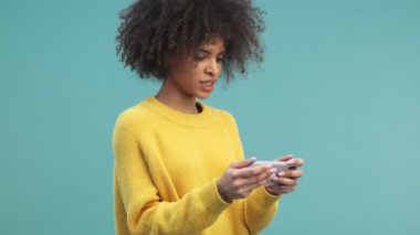 Kafası karışmış kıvırcık saçlı Afro-Amerikan bir kadın akıllı telefonundan garip bir şey izliyor ve stüdyodaki mavi duvarın arkasında tek başına duruyor.