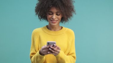Gülümseyen, kıvırcık saçlı Afro-Amerikan bir kadın stüdyoda mavi duvar arkasında tek başına duran akıllı telefonuna yazı yazıyor.