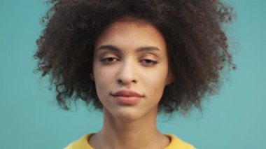 Kıvırcık saçlı mutlu Afro-Amerikan bir kadının yakın çekim görüntüsü gözlerini açıyor ve stüdyoda mavi duvarın arkasında tek başına duran kameraya bakıyor.