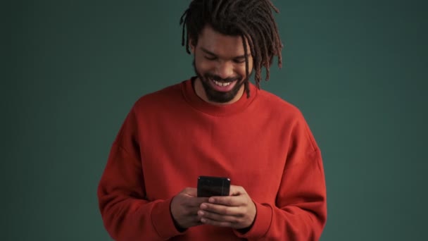 一个笑着的年轻的非洲裔美国人正在工作室里用他的智能手机站在绿墙后面 — 图库视频影像