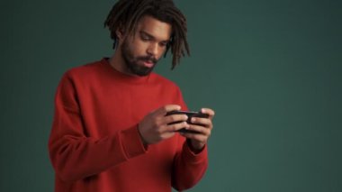 Gülümseyen Afrikalı genç bir Amerikalı akıllı telefonuyla oyun oynuyor. Stüdyoda yeşil duvar arkasında tek başına duruyor.