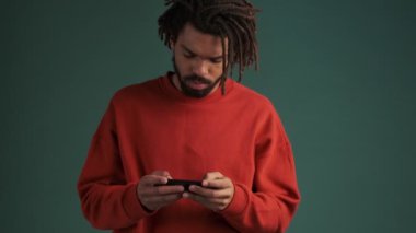 Odaklanmış Afro-Amerikan bir adam akıllı telefonuyla oyun oynuyor ve stüdyoda yeşil duvar arkasında tek başına duran başarılı bir jest yapıyor.