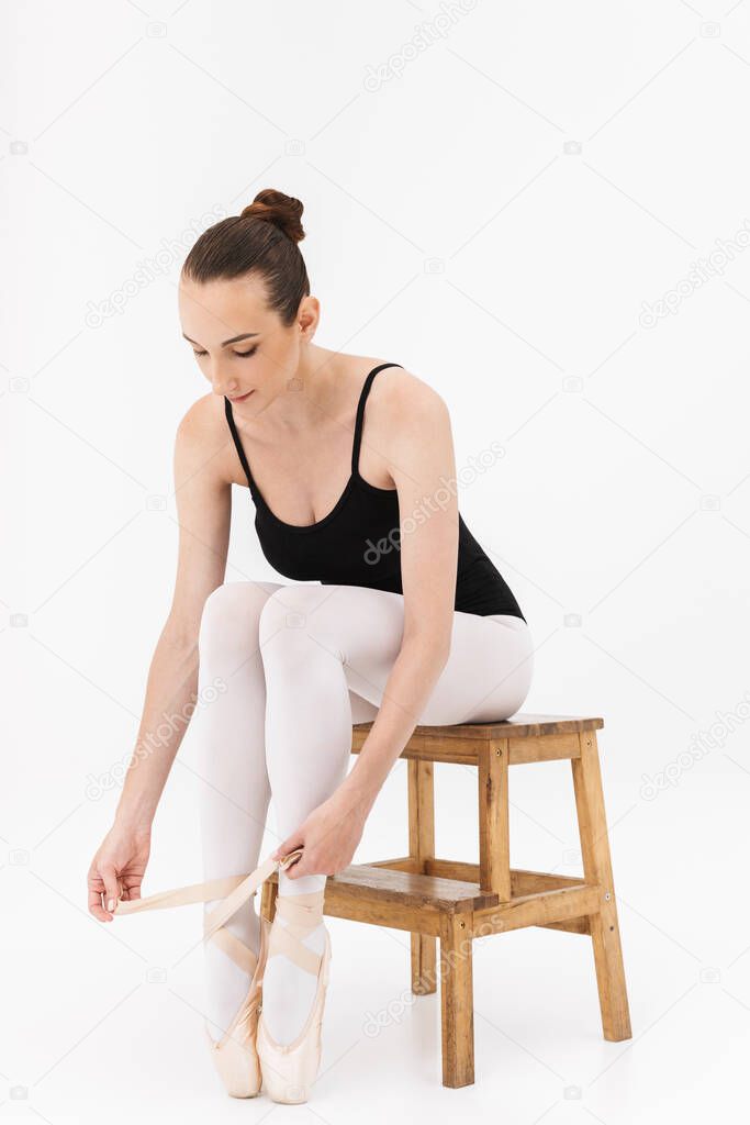 Image of charming young woman ballerina sitting on chair and wearing pointe shoes isolated over white wall background