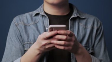 Genç Asyalı bir adamın kırpılmış görüntüsü akıllı telefonuyla stüdyodaki koyu mavi duvar arkaplanının üzerinde yazıyor.
