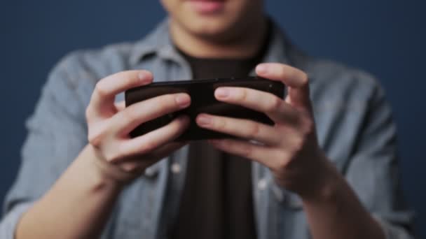 若いアジア人男性のクロップドビューは スタジオでダークブルーの壁の背景に隔離されたそれにゲームをプレイしながら 水平方向に彼のスマートフォンを保持しています — ストック動画