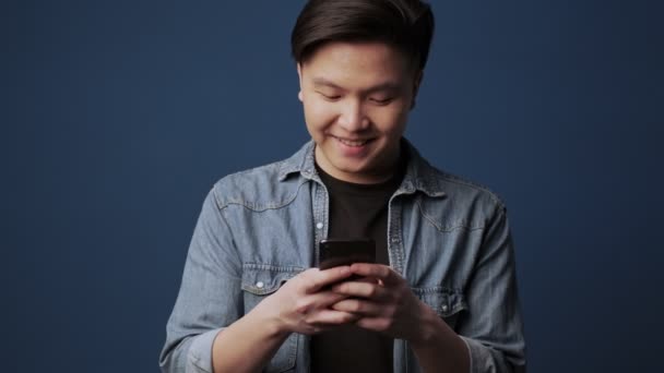 幸せな笑顔の若いアジア人男性はスタジオで暗い青色の壁の背景に隔離された彼のスマートフォンを使用しています — ストック動画