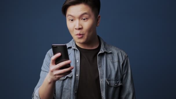 驚くべき若いアジアの男は彼のスマートフォンを探しているとタイプそれに隔離されていますダークブルーの壁の背景の上にスタジオ — ストック動画