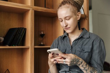 Rastalı ve piercingli genç bir kızın cep telefonu kullanarak rafın yanında duruşu..