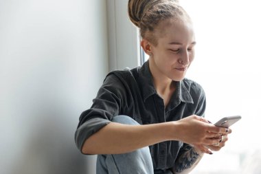 Pencerenin yanında cep telefonu kullanan, rastalı ve piercingli mutlu genç bir kızın resmi.