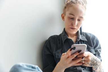 Pencerenin yanında cep telefonu kullanan, rastalı ve piercingli genç bir kız resmi.