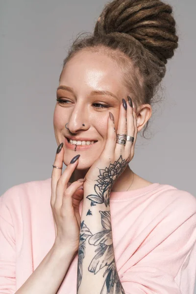 Image of happy extraordinary girl with tattoo and face piercing touching her face isolated over gray background