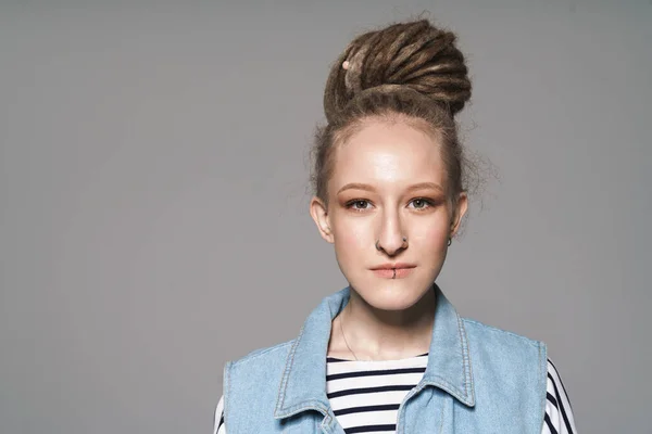 Image of young extraordinary girl with face piercing and dreadlocks looking at camera isolated over gray background
