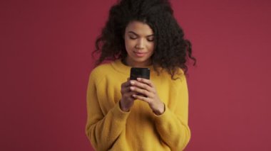 Genç Afrikalı pozitif kadın koyu kırmızı bordo arka planda cep telefonu kullanarak izole edilmiş.