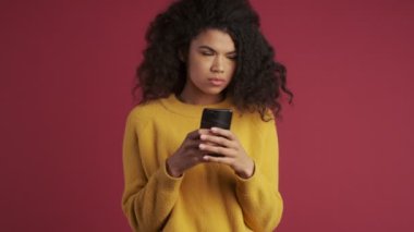 Genç Afrikalı hoşnutsuz kadın cep telefonu kullanarak koyu kırmızı bordo arka planda izole edilmiş.