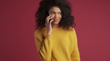 Kafası karışmış genç Afrikalı bir kadın koyu kırmızı bordo arka plandan izole edilmiş cep telefonuyla konuşuyor.