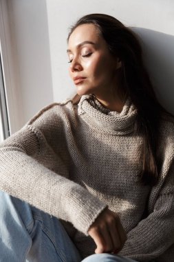 Pencerenin pervazında otururken gözleri kapalı poz veren rahat Asyalı kadın resmi