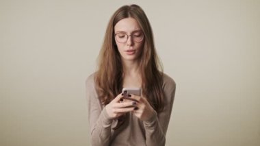 Gri duvar arkaplanı üzerinde cep telefonu kullanarak izole edilmiş güzel, konsantre olmuş genç ve güzel bir kadın.