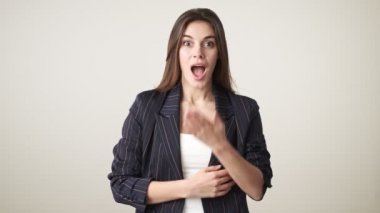 Gündelik kıyafetler giyen mutlu genç bir kadın, stüdyoda beyaz arka planda tek başına ayakta alkışlıyor ve seviniyor.