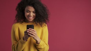 Genç Afrikalı pozitif kadın koyu kırmızı bordo arka planda cep telefonu kullanarak izole edilmiş.
