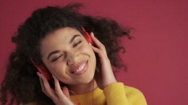 Genç Afrikalı pozitif kadın koyu kırmızı bordo arka planda izole edilmiş. Kulaklıkla müzik dinliyor.