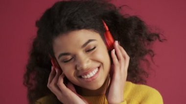 Genç Afrikalı mutlu kadın koyu kırmızı bordo arka planda izole edilmiş kulaklıkla müzik dinliyor.