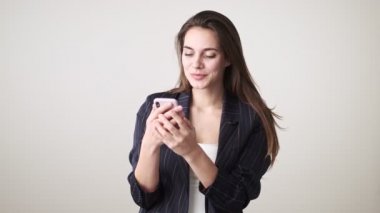 Gündelik giysiler giyen güzel genç bir kadın, atölyedeki beyaz arka planda tek başına duran akıllı telefonunu kullanıyor.