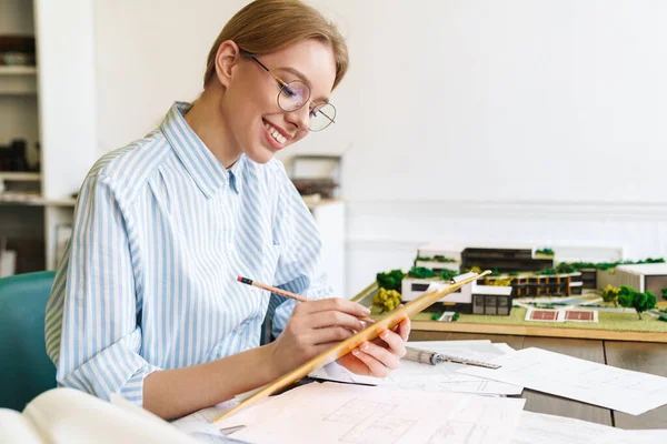 Photo of joyful young woman architect in eyeglasses working with drawings while designing draft at workplace